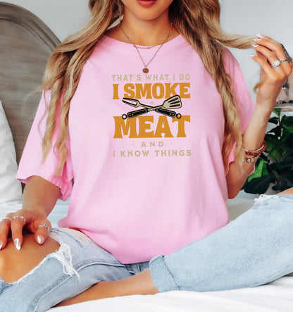 That's What I Do, I Smoke Meat & I Know Things T-shirt featuring bold text in yellow and white with crossed grilling tools at the center. Printed on a blossom pink unisex tee, this fun BBQ-themed design is perfect for grill masters and smoking enthusiasts. Ideal for Father’s Day, cookouts, and casual wear for those proud of their grilling skills and witty sense of humor.