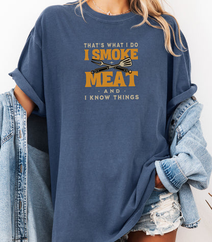 That is What I Do, I Smoke Meat & I Know Things T-shirt featuring bold text in yellow and white with crossed grilling tools at the center. Printed on a denim unisex tee, this fun BBQ-themed design is perfect for grill masters and smoking enthusiasts. Ideal for Fathers Day, cookouts, and casual wear for those proud of their grilling skills and witty sense of humor.