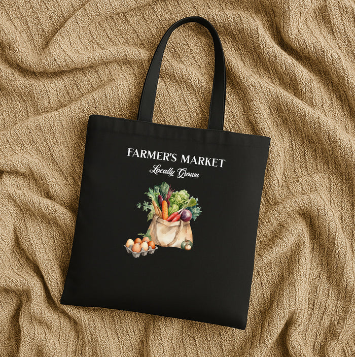 A black cotton canvas tote bag with 'FARMER'S MARKET' text and an illustration of vegetables and fruits on the front laying flat on a tan fabric background.