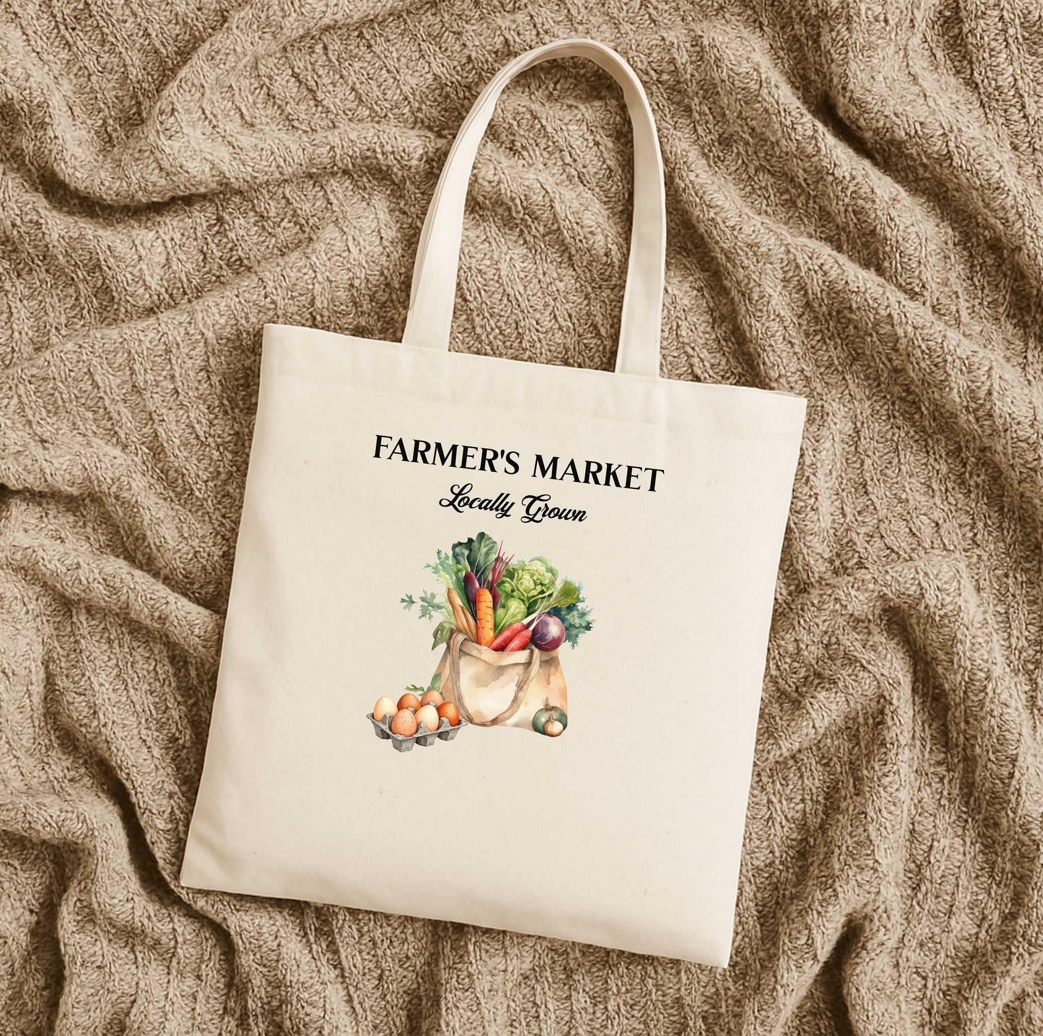 A natural cotton canvas tote bag with 'FARMER'S MARKET' text and an illustration of vegetables and fruits on the front laying flat on a tan fabric.