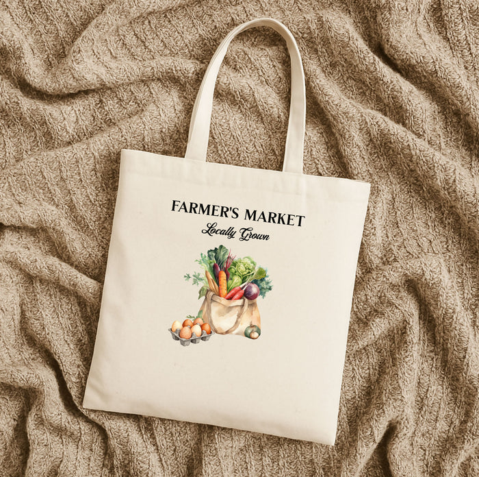 A natural cotton canvas tote bag with 'FARMER'S MARKET' text and an illustration of vegetables and fruits on the front laying flat on a tan fabric.