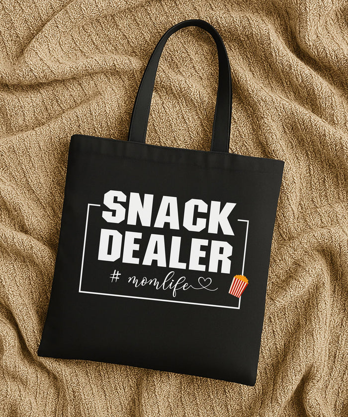 Black tote bag with 'SNACK DEALER #momlife' text laying flat on a tan fabric background