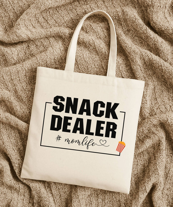 A Natural Tote bag with 'Snack Dealer #momlife' text laying flat on a tan textured fabric background