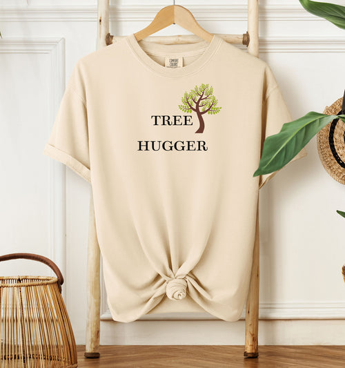 Tree Hugger T-shirt featuring a simple nature-inspired design with a brown and green tree graphic placed between the words Tree and Hugger in classic black font. Printed on a ivory unisex Comfort Colors tee, this eco-friendly design is perfect for environmentalists, outdoor lovers, hikers, and anyone passionate about protecting nature and forests.
