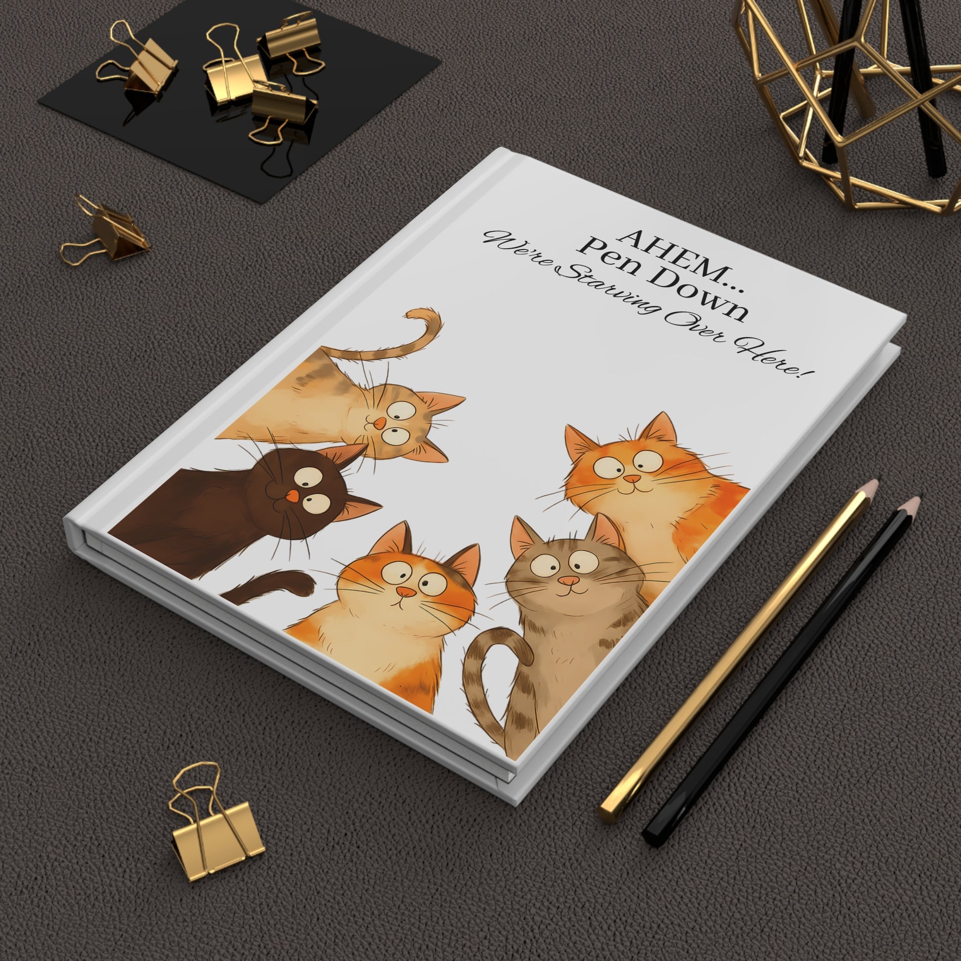 Hardcover journal with cartoon cats on the cover, surrounded by office supplies on a dark surface
