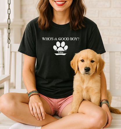Woman wearing a black t-shirt that reads “Who’s a Good Boy?” above a white paw print graphic with the name “Toby”.  Sitting cross-legged with a golden retriever puppy, the shirt is a playful and heartwarming tribute to a beloved pet—perfect for dog moms and puppy lovers.