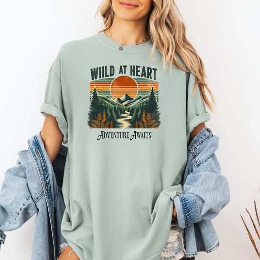 Vintage-style “Wild at Heart” graphic featuring a bold retro sunset over a pine forest and mountain scene, with a winding river flowing through the center. Warm earth tones—orange, green, and cream—create a nostalgic outdoor vibe. Ideal for adventure lovers and nature enthusiasts on a Comfort Colors Bay (light green) tee.
