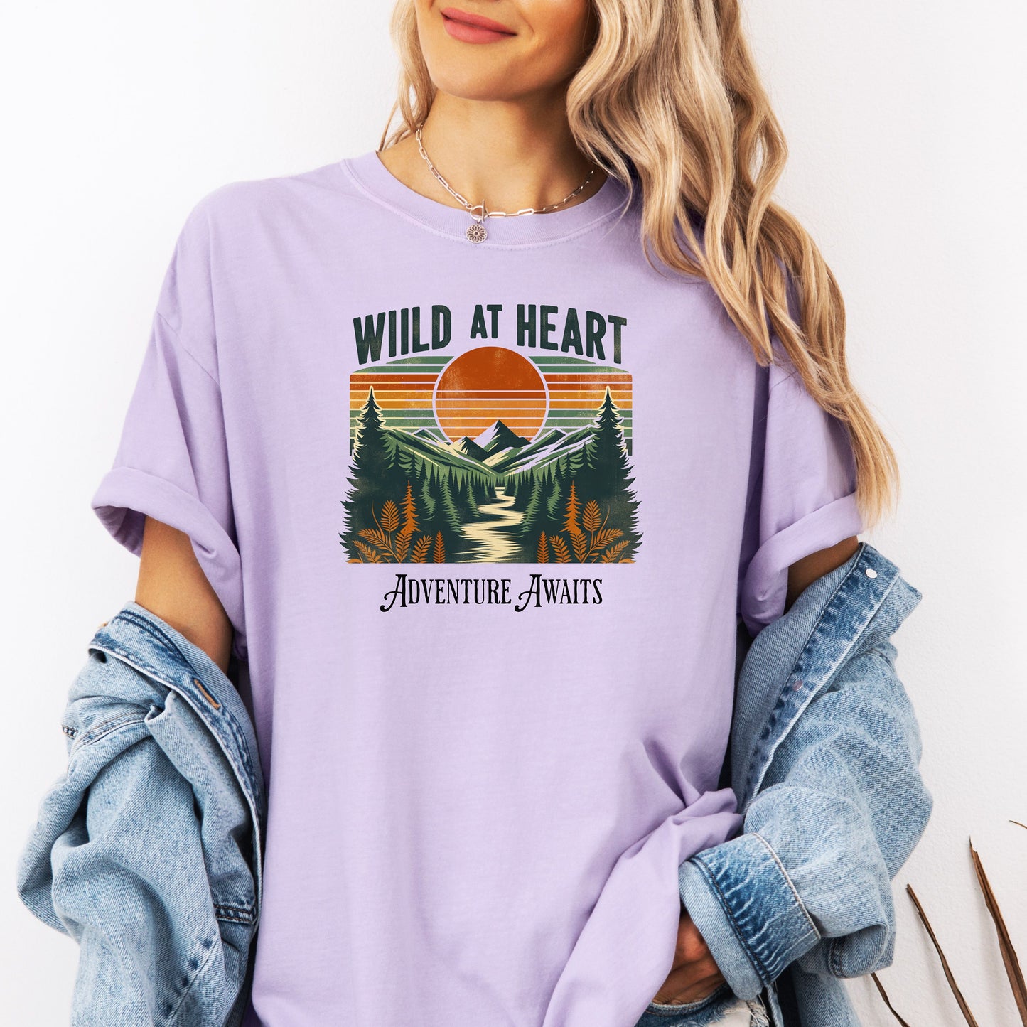 Vintage-style “Wild at Heart” graphic featuring a bold retro sunset over a pine forest and mountain scene, with a winding river flowing through the center. Warm earth tones—orange, green, and cream—create a nostalgic outdoor vibe. Ideal for adventure lovers and nature enthusiasts on a Comfort Colors Orchid T-shirt.