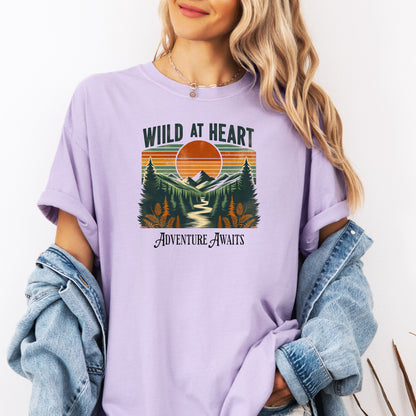 Vintage-style “Wild at Heart” graphic featuring a bold retro sunset over a pine forest and mountain scene, with a winding river flowing through the center. Warm earth tones—orange, green, and cream—create a nostalgic outdoor vibe. Ideal for adventure lovers and nature enthusiasts on a Comfort Colors Orchid T-shirt.