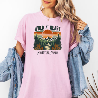 Vintage-style “Wild at Heart” graphic featuring a bold retro sunset over a pine forest and mountain scene, with a winding river flowing through the center. Warm earth tones—orange, green, and cream—create a nostalgic outdoor vibe. Ideal for adventure lovers and nature enthusiasts on a Comfort Colors Blossom (pink) tee.