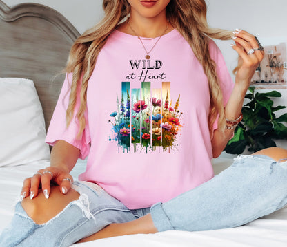 Blossom pink “Wild at Heart” T-shirt featuring a vibrant watercolor-style design of wildflowers in pink, red, purple, yellow, and blue, set against vertical gradient bars in teal, green, and warm orange tones. The text “Wild at Heart” appears above the floral artwork in elegant black lettering. Perfect for nature lovers and free spirits, this casual tee offers a relaxed, stylish fit ideal for everyday wear.