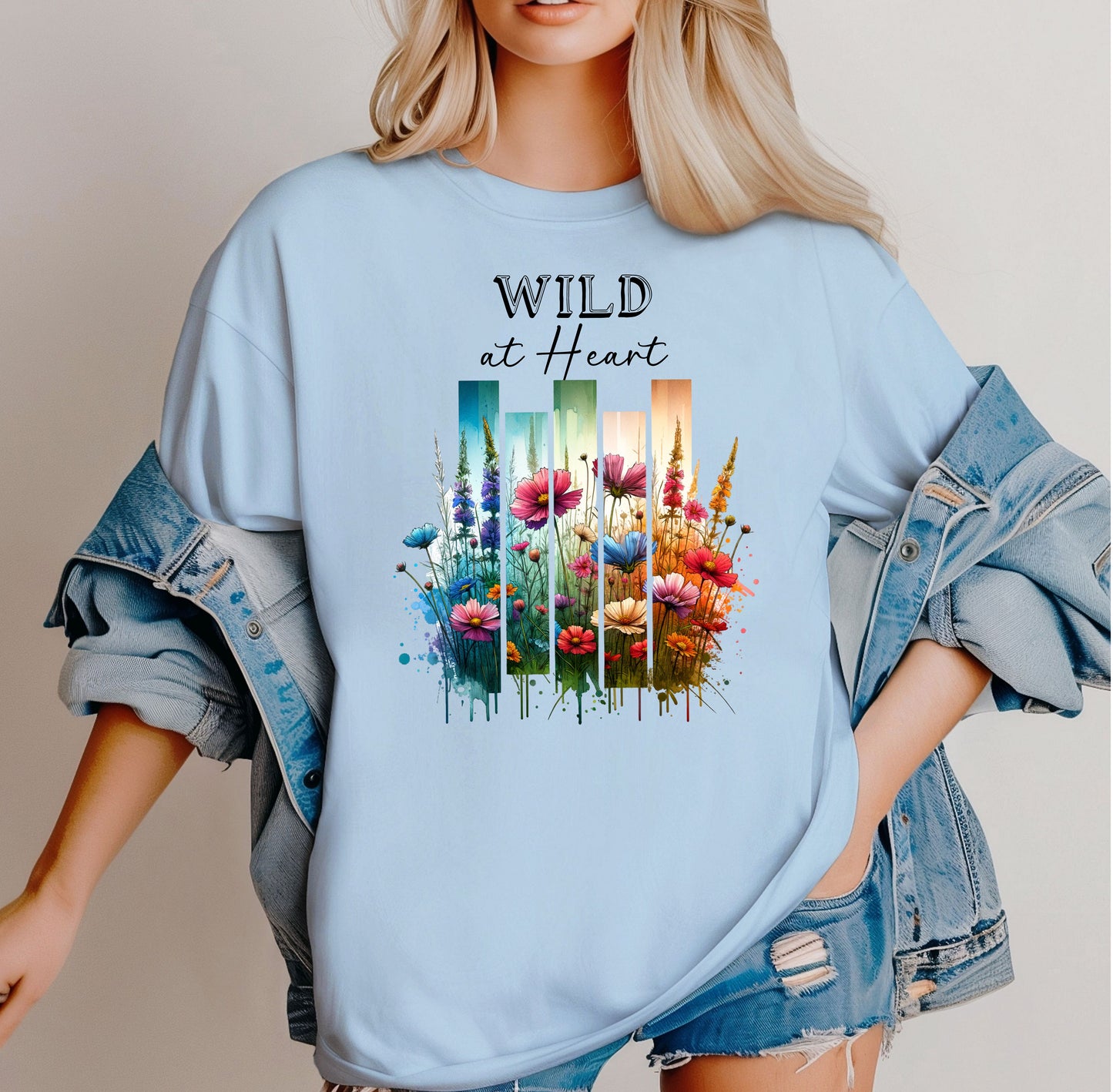 Chambray light blue “Wild at Heart” T-shirt featuring a vibrant watercolor-style design of wildflowers in pink, red, purple, yellow, and blue, set against vertical gradient bars in teal, green, and warm orange tones. The text “Wild at Heart” appears above the floral artwork in elegant black lettering. Perfect for nature lovers and free spirits, this casual tee offers a relaxed, stylish fit ideal for everyday wear.