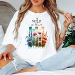 White “Wild at Heart” T-shirt featuring a vibrant watercolor-style design of wildflowers in pink, red, purple, yellow, and blue, set against vertical gradient bars in teal, green, and warm orange tones. The text “Wild at Heart” appears above the floral artwork in elegant black lettering. Perfect for nature lovers and free spirits, this casual tee offers a relaxed, stylish fit ideal for everyday wear.