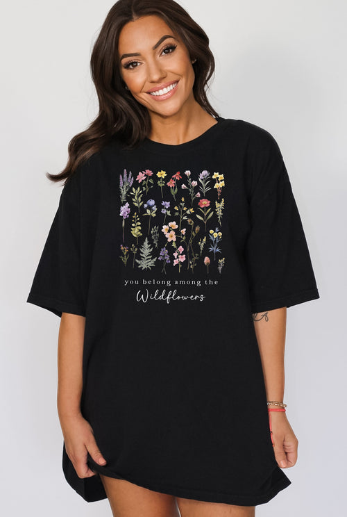 A woman standing wearing the black t-shirt.  The text reads 'you belong among the Wildflowers'.  Above the text shows a grid of wildflowers.