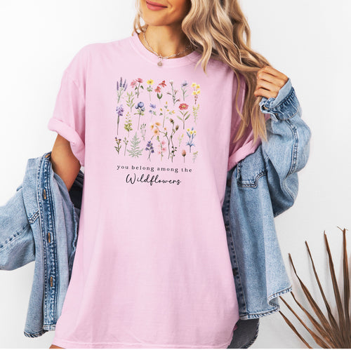 A woman standing wearing the blossom pink t-shirt.  The text reads 'you belong among the Wildflowers'.  Above the text shows a grid of wildflowers.