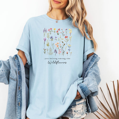 A woman standing wearing the chambray light blue t-shirt.  The text reads 'you belong among the Wildflowers'.  Above the text shows a grid of wildflowers.