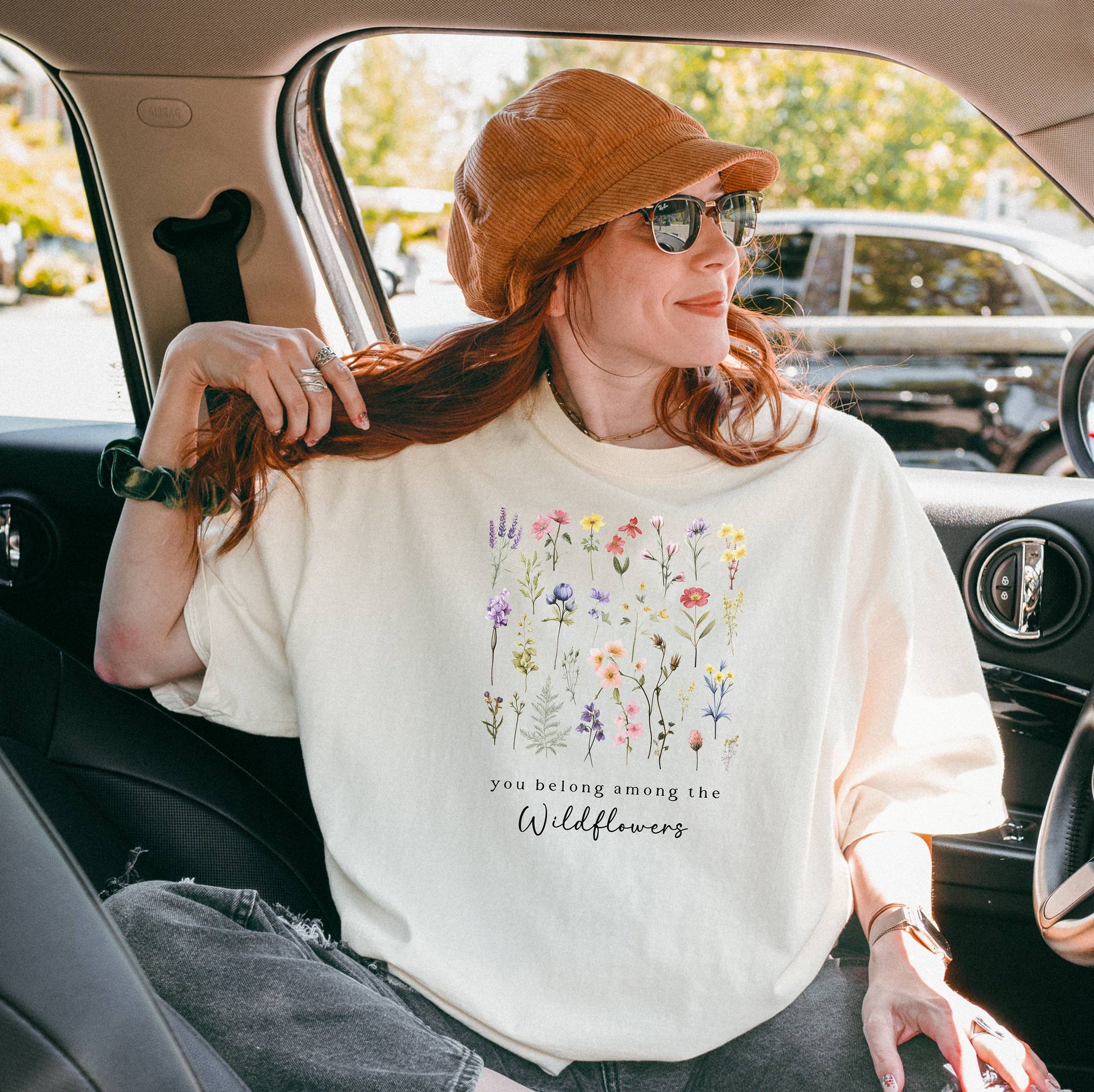 A girl sitting in a car wearing the ivory t-shirt.  The text reads 'you belong among the Wildflowers'.  Above the text shows a grid of wildflowers.