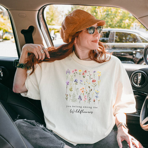 A girl sitting in a car wearing the ivory t-shirt.  The text reads 'you belong among the Wildflowers'.  Above the text shows a grid of wildflowers.