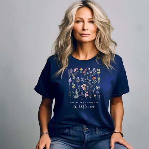 A woman sitting wearing the true navy t-shirt.  The text reads 'you belong among the Wildflowers'.  Above the text shows a grid of wildflowers.