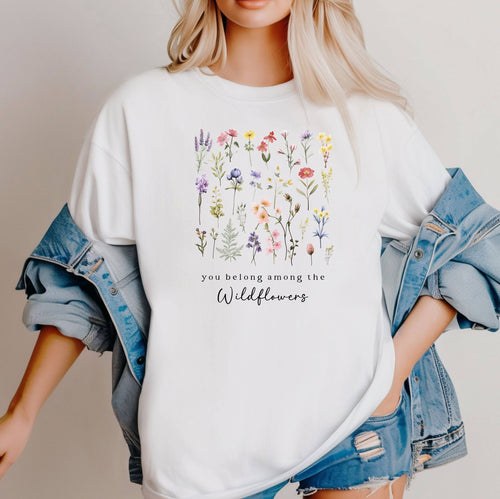 A girl standing wearing the white t-shirt.  The text reads 'you belong among the Wildflowers'.  Above the text shows a grid of wildflowers.