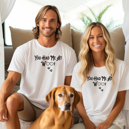 Smiling man and woman sitting indoors with a brown dog, both wearing matching white T-shirts featuring the phrase “You Had Me At Woof” in playful black text. The design includes heart shapes inside the letters and two small paw prints beneath, creating a cute and stylish look perfect for dog lovers and pet owners.