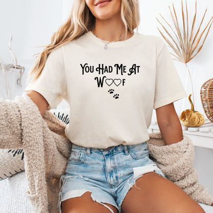 Person wearing a soft cream T-shirts featuring the phrase “You Had Me At Woof” in playful black text. The design includes heart shapes inside the letters and two small paw prints beneath, creating a cute and stylish look perfect for dog lovers and pet owners.