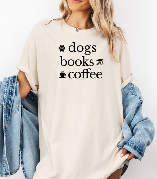 Black graphic design on an Ivory t-shirt featuring the phrase “Dogs, Books, Coffee” in bold, playful lettering. Decorative icons of a dog paw, stacked books, and a steaming coffee cup enhance the cozy, laid-back vibe—perfect for dog lovers, bookworms, and caffeine enthusiasts alike.